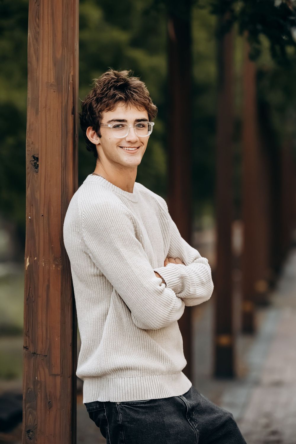 Young man in beige sweater and clear glasses leaning against wooden post with arms crossed, smiling outdoors.