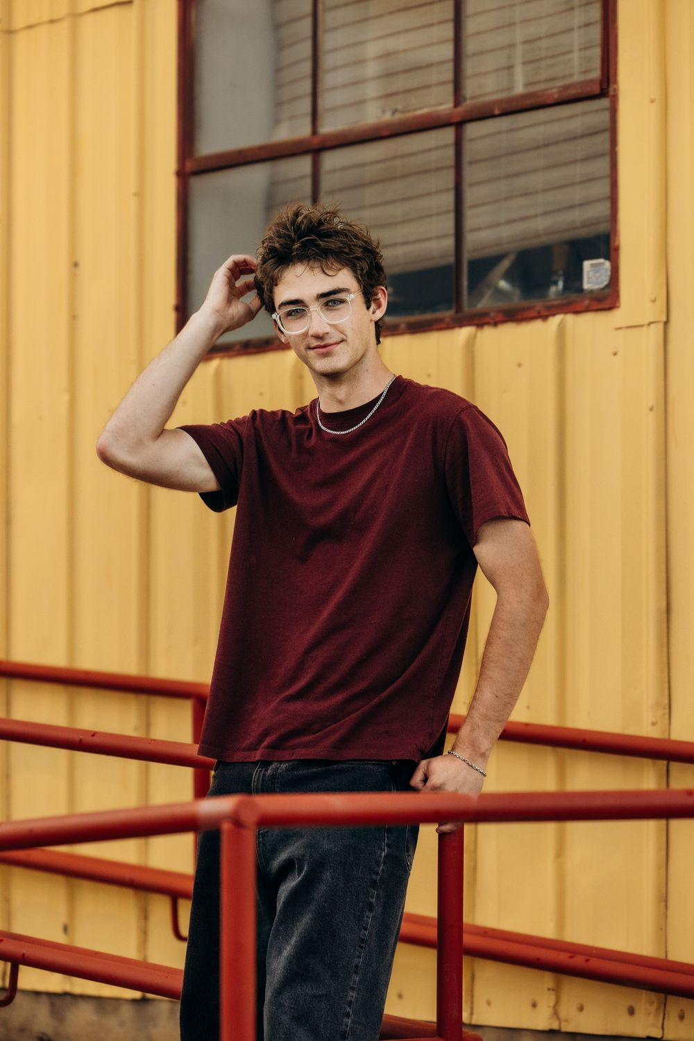 Young man in maroon t-shirt and glasses poses confidently against a yellow industrial building with red railings.