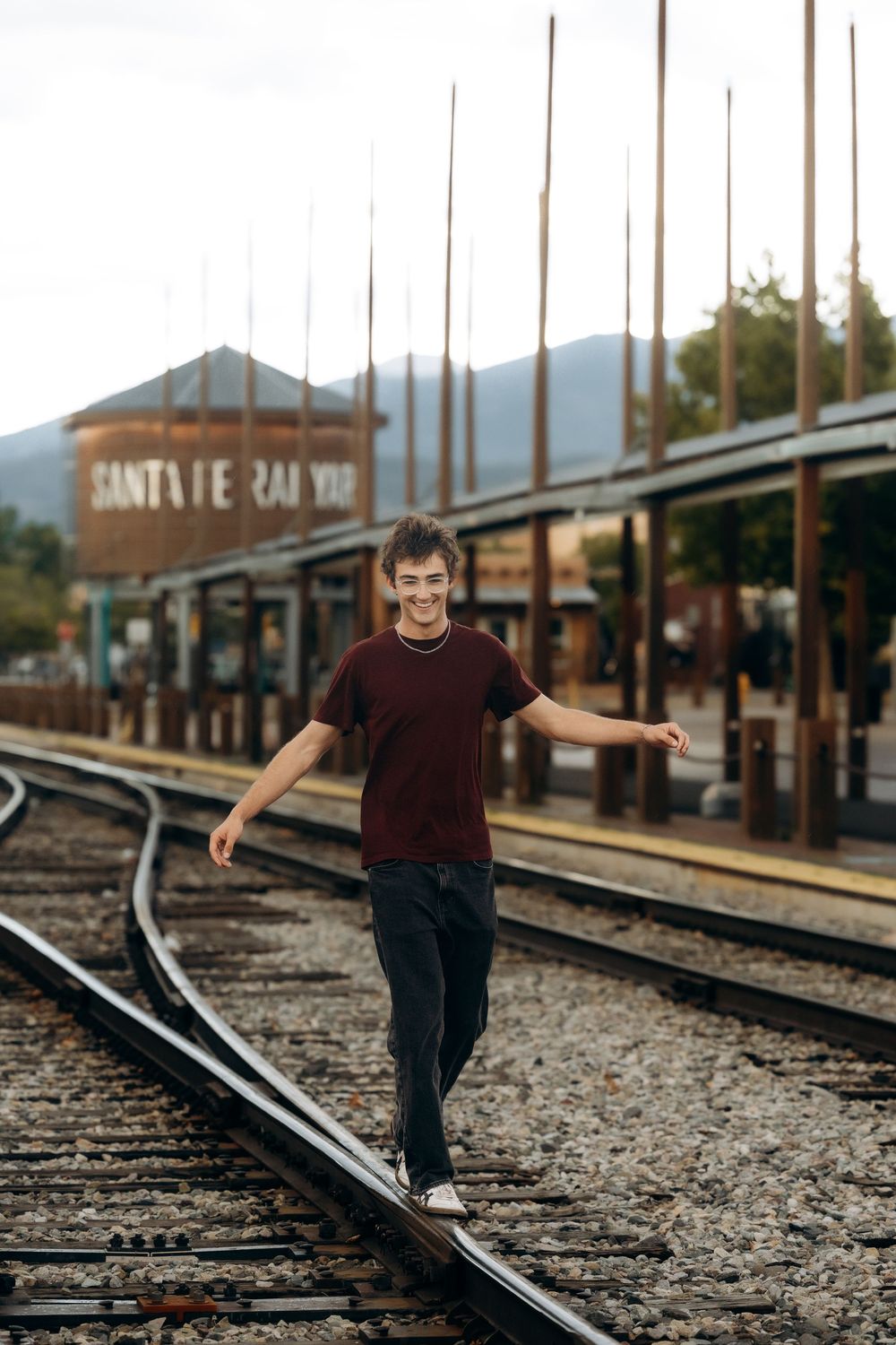 A smiling young man balances on railroad tracks at Santa Fe Railway station, arms outstretched, wearing a maroon t-shirt.