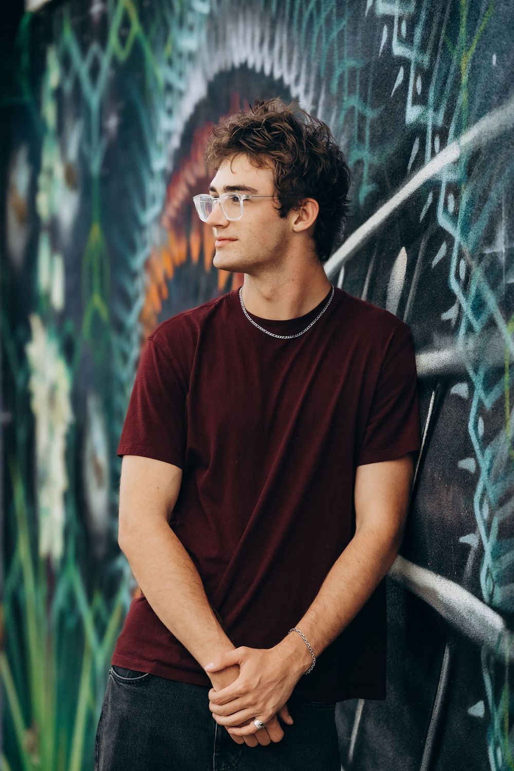 Young man with glasses in a maroon t-shirt leaning against a colorful graffiti wall with hands clasped.