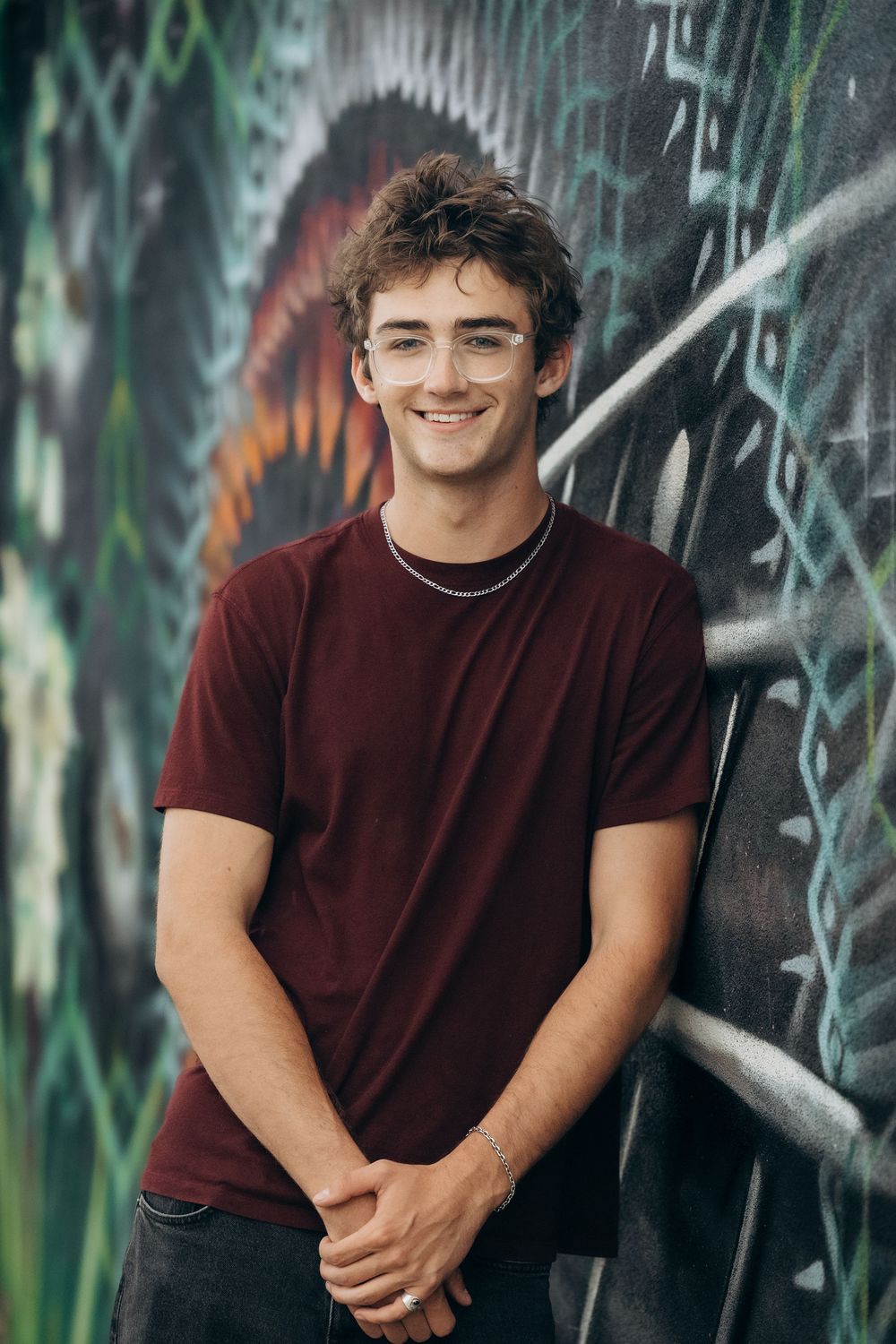 Smiling young man in a maroon t-shirt leaning against a colorful graffiti wall with hands clasped.