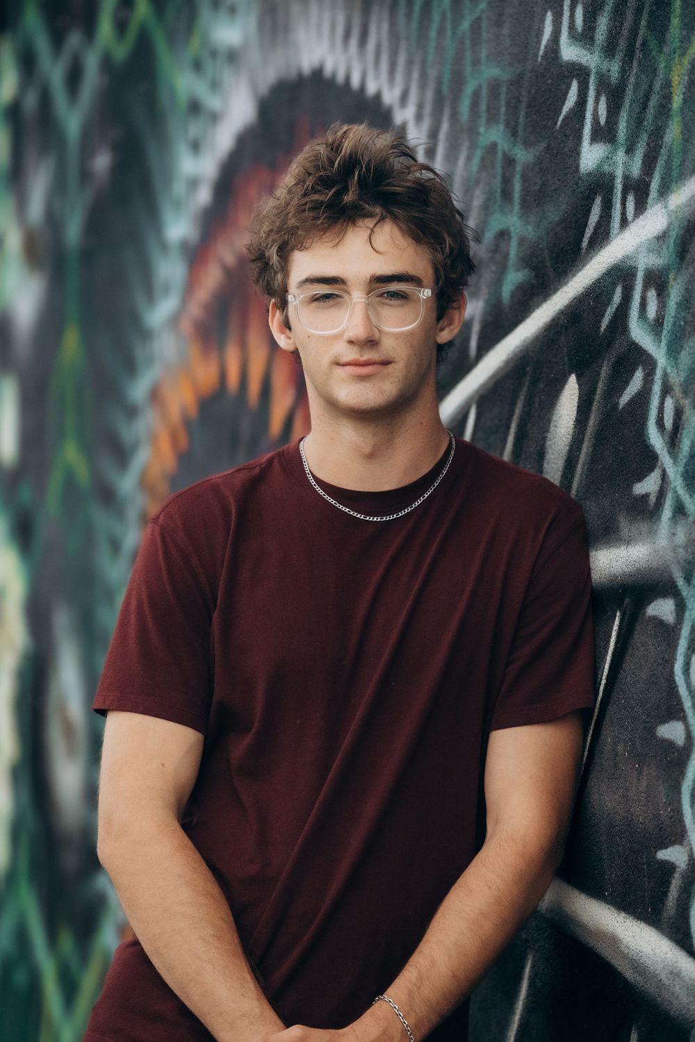 Young man in maroon t-shirt and clear glasses leaning against colorful graffiti wall.