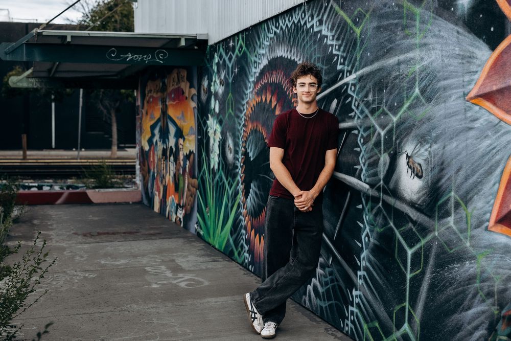 Young man in red t-shirt posing against colorful graffiti mural wall in urban outdoor setting.