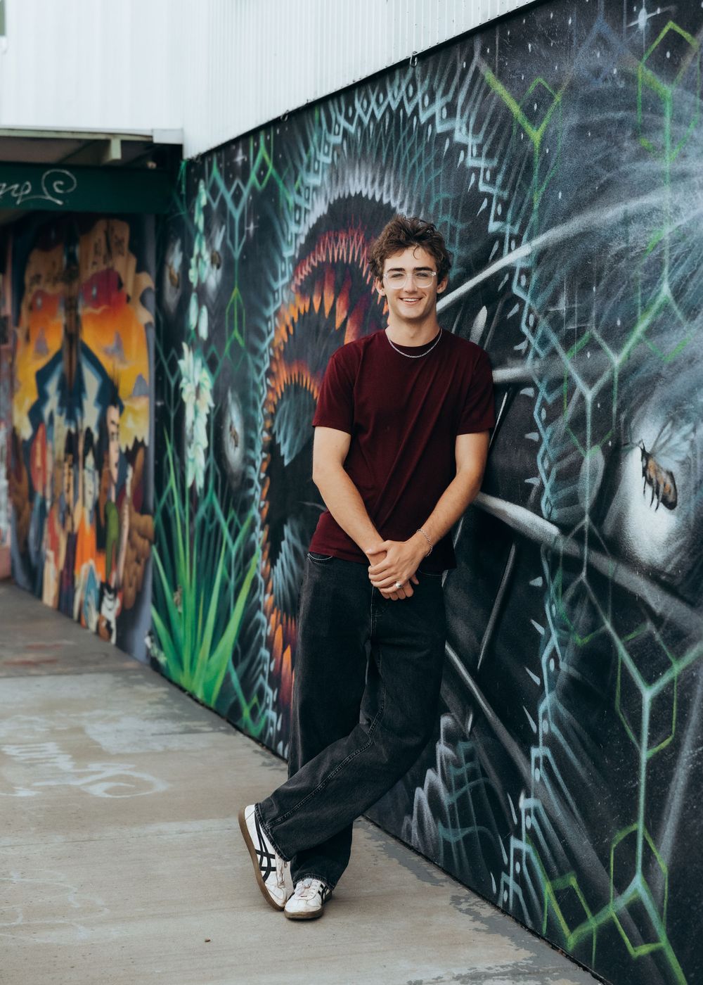 Young man in red shirt and black jeans leaning against a vibrant street art mural wall, smiling casually.