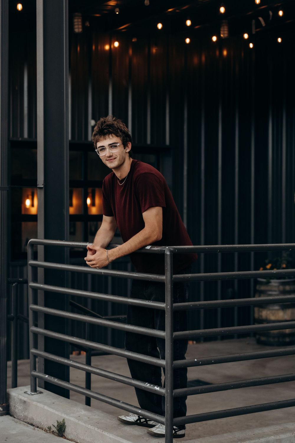 Young man in burgundy shirt leaning on metal railing, smiling outdoors with warm string lights in background.