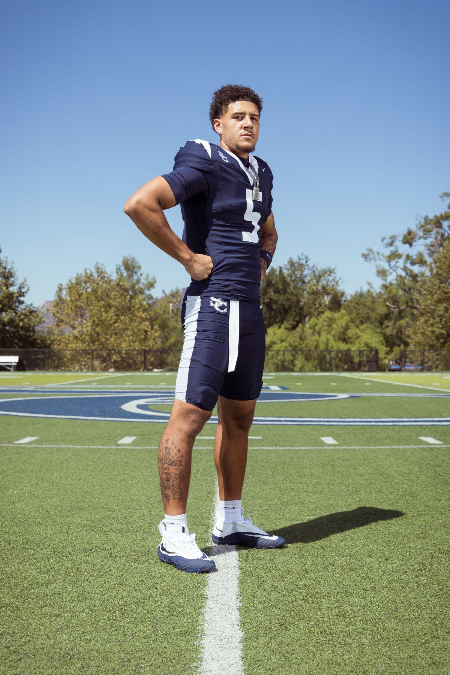 Football player in navy uniform stands confidently on green field under clear blue sky.