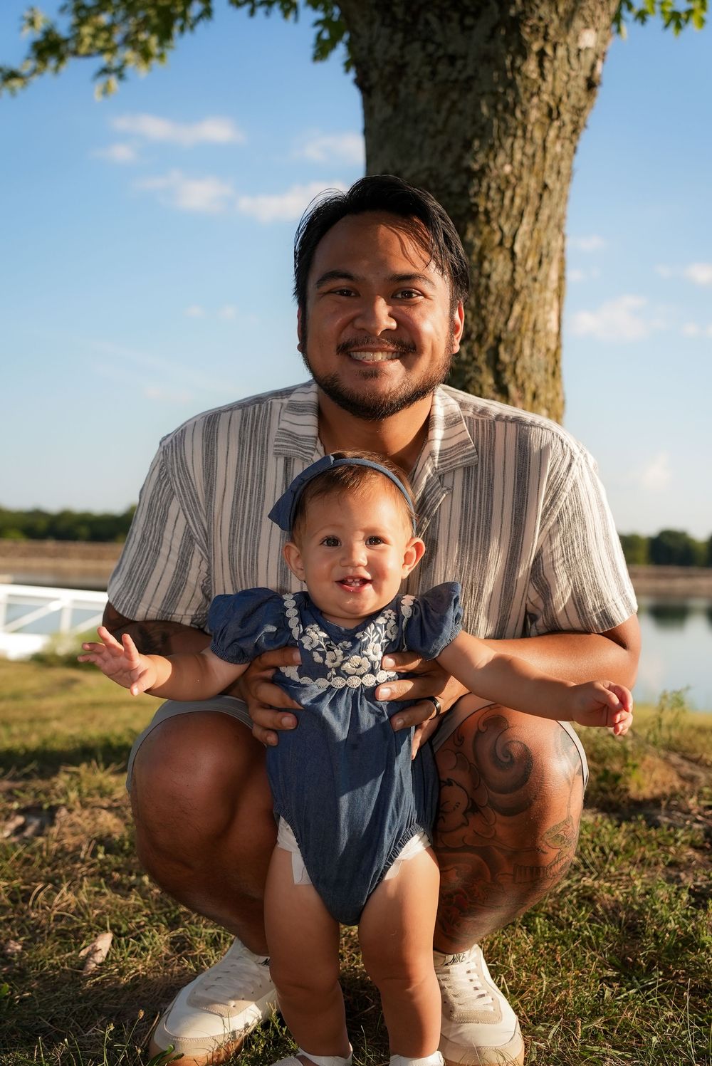 Smiling man crouching outdoors holding a happy baby girl in a blue outfit near a tree by a lake.