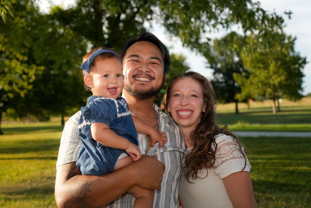 Happy family of three smiling outdoors in a sunny green park, parents holding a cheerful baby girl.