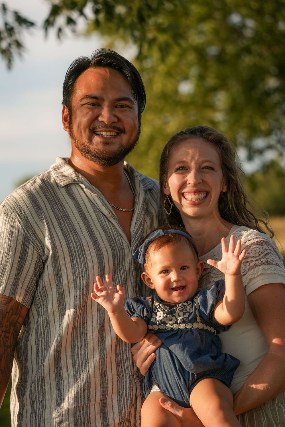 Happy family portrait outdoors with smiling parents holding a cheerful baby girl waving at the camera in golden sunlight.