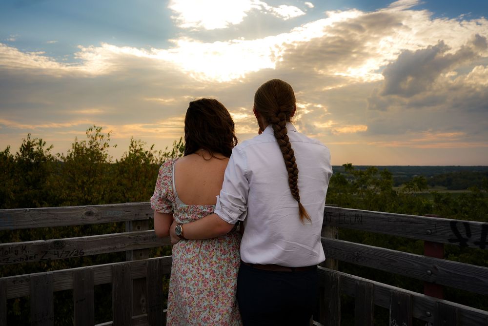 Couple standing on wooden deck watching stunning sunset over green landscape, viewed from behind.