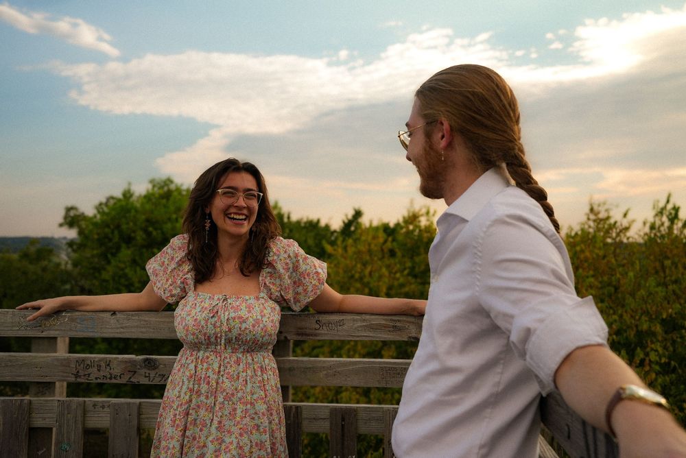 A laughing woman in a floral dress leans on a wooden railing while a man in white shirt faces her at sunset.