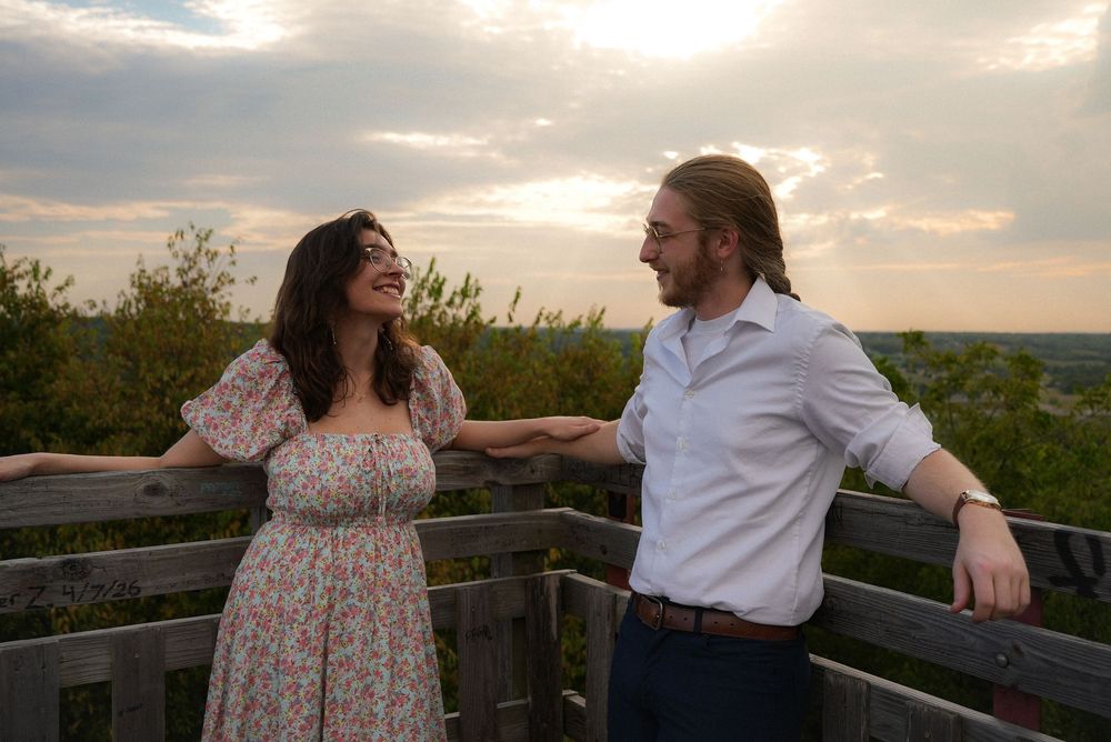 A happy couple smiles at each other while leaning on a wooden railing with a scenic cloudy sky backdrop.