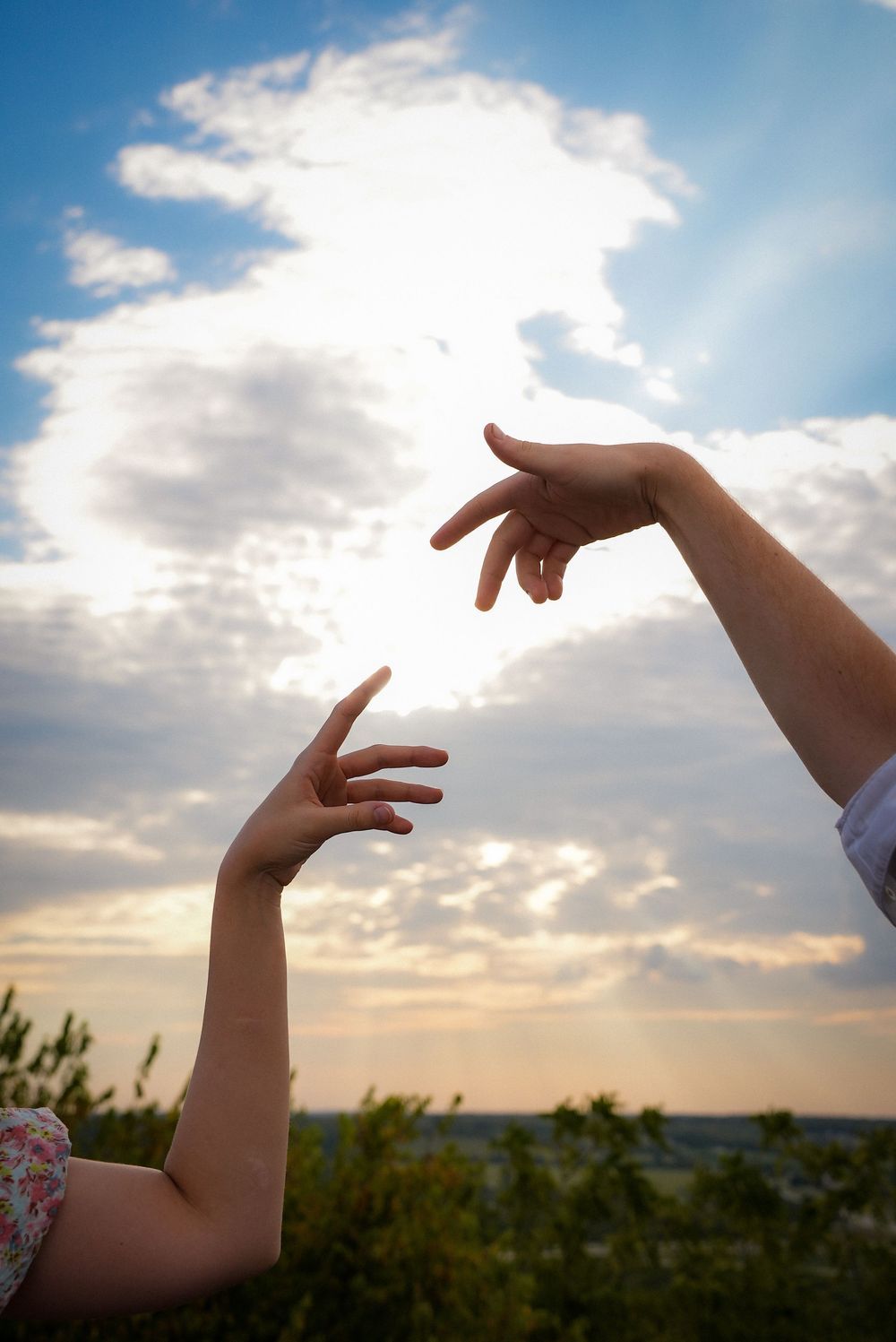 Two hands reaching toward each other against a dramatic sunset sky with clouds.