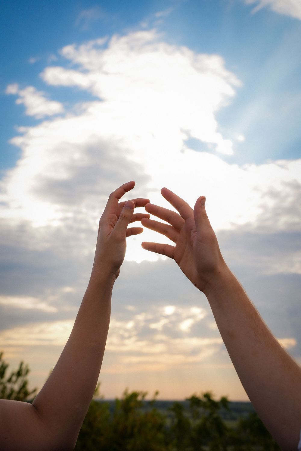 Two hands reach upward toward a dramatic cloudy sky with sunlight breaking through at golden hour.