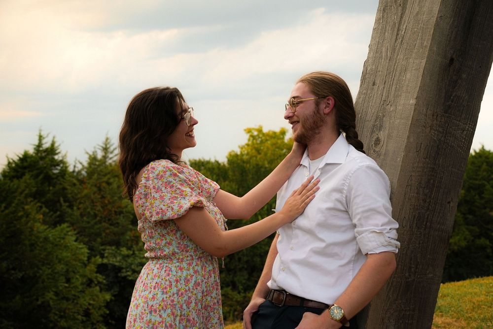A couple shares a tender moment outdoors, woman in floral dress touching mans face near a wooden post.