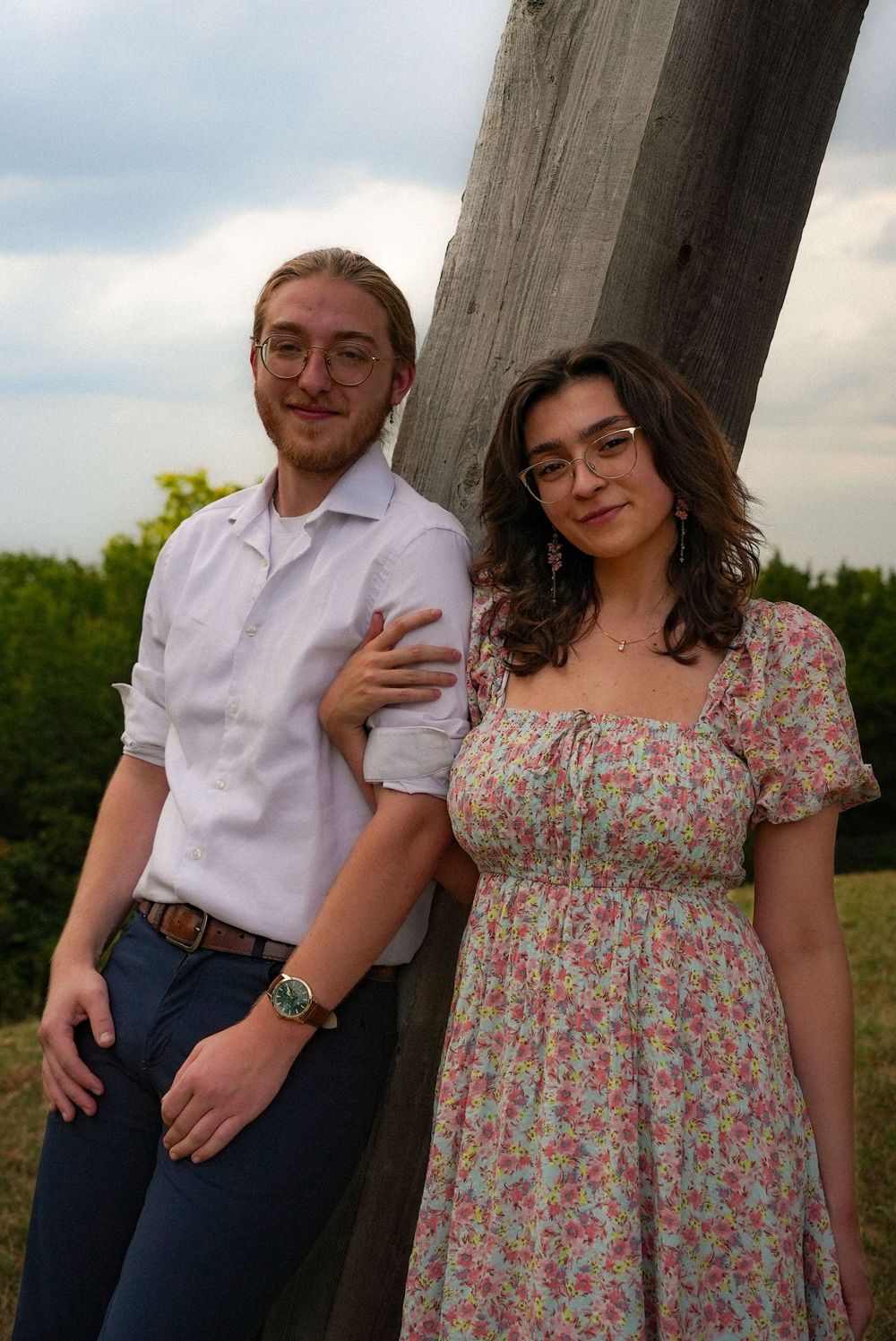 A young couple poses together leaning against a wooden post outdoors with a cloudy sky background.
