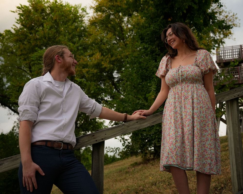A couple holds hands while standing on wooden steps surrounded by lush green trees at dusk.