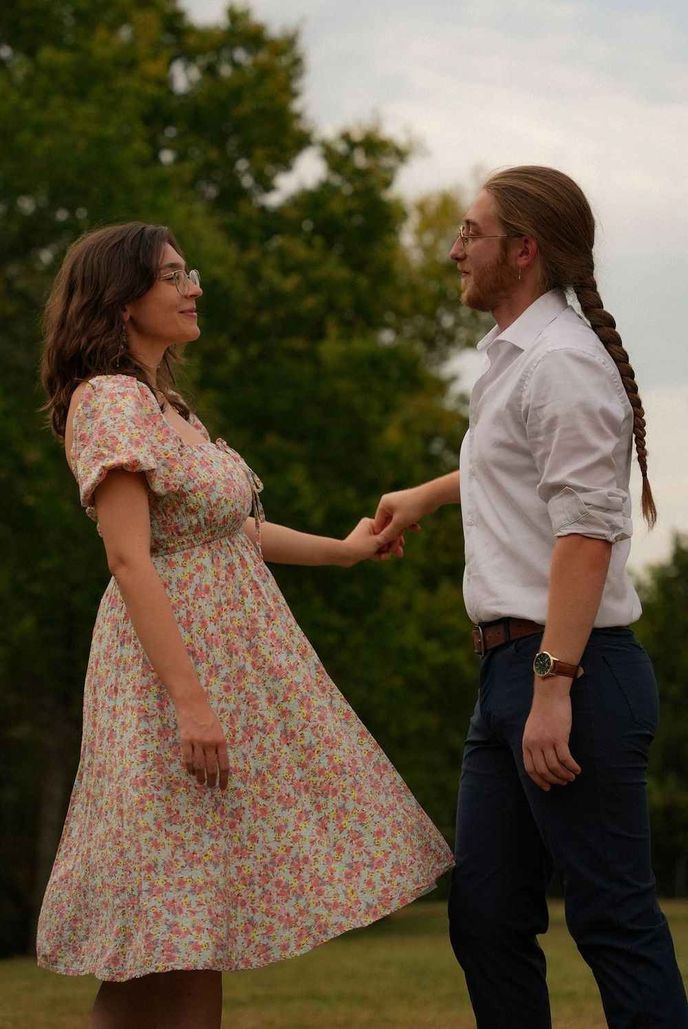 Couple holding hands in a park, woman in floral dress and man with braided hair in white shirt.