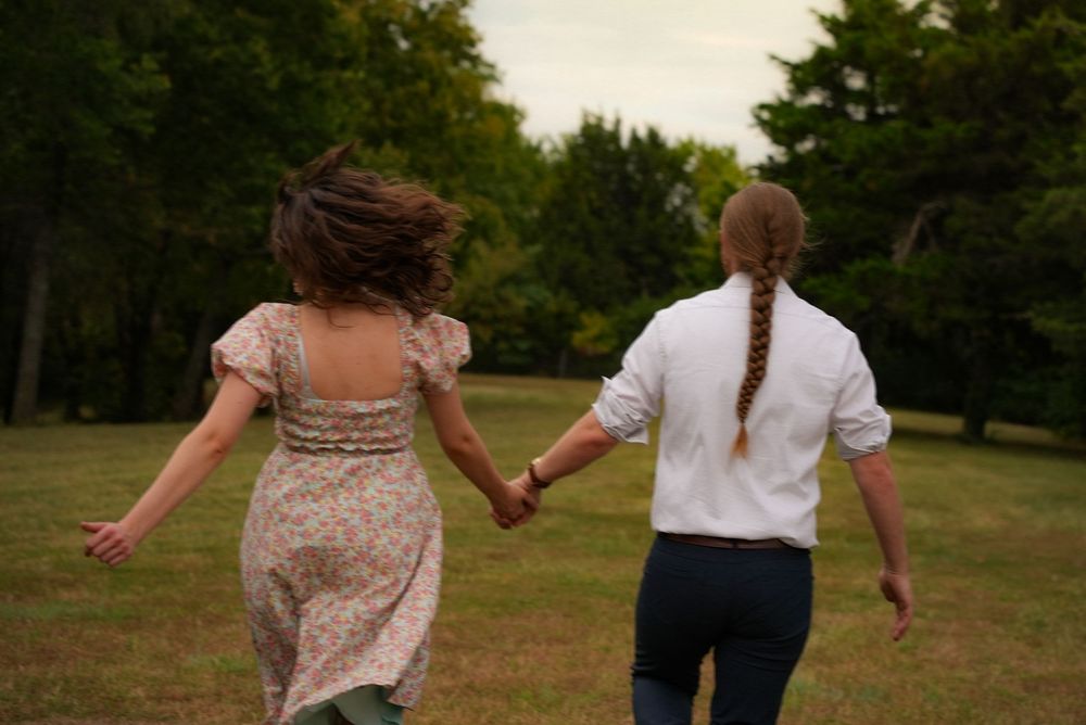 Two women holding hands running through a green park, one in a floral dress and one in a white shirt with a braid.