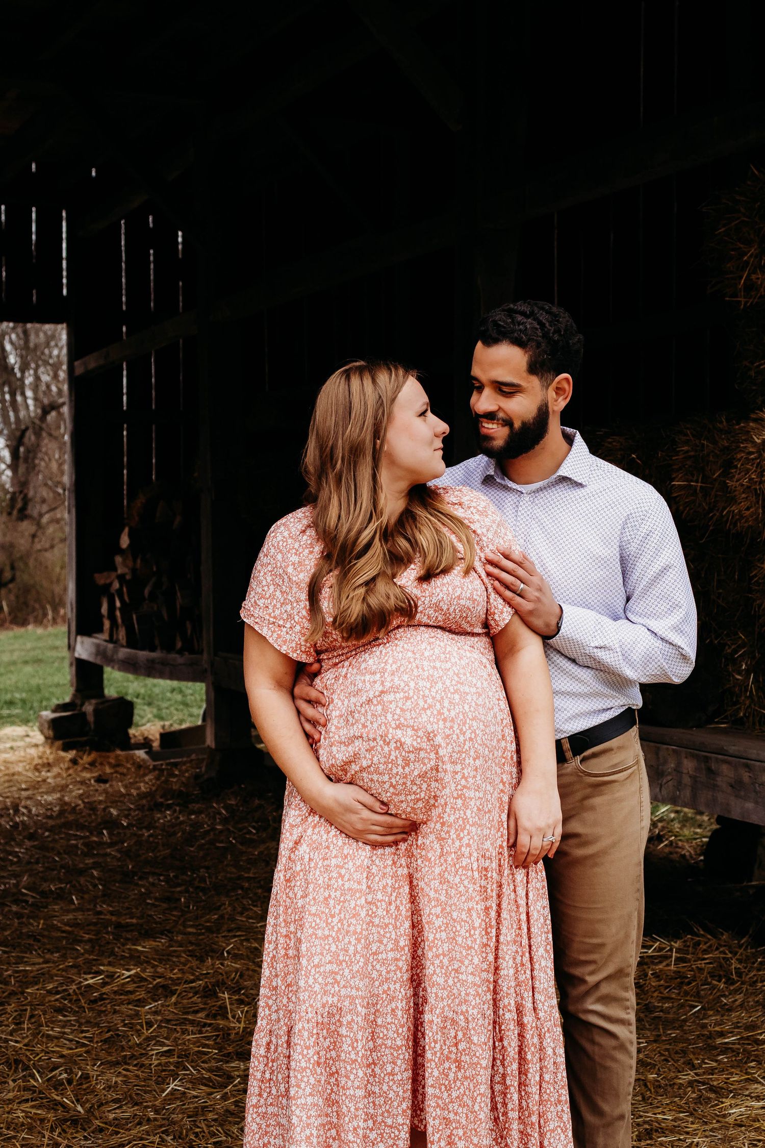 A couple shares a tender moment in a rustic barn setting, with the woman wearing a flowing pink floral maternity dress.