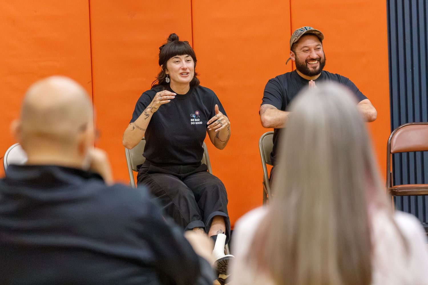 Two people seated in chairs speak to an audience in front of an orange wall during a panel discussion.