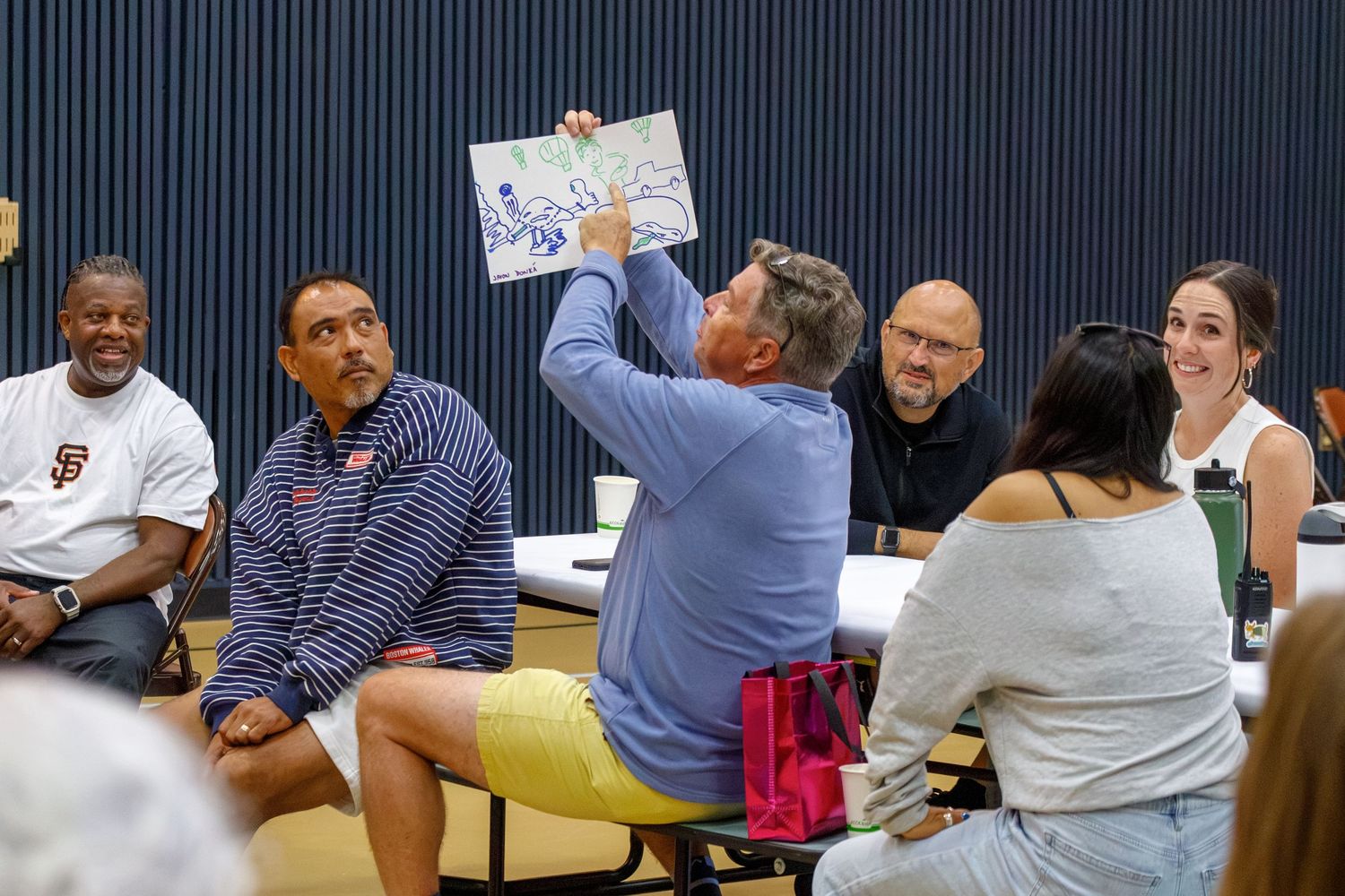 Group of adults in a meeting room, one person holding up a colorful map or artwork for others to view.