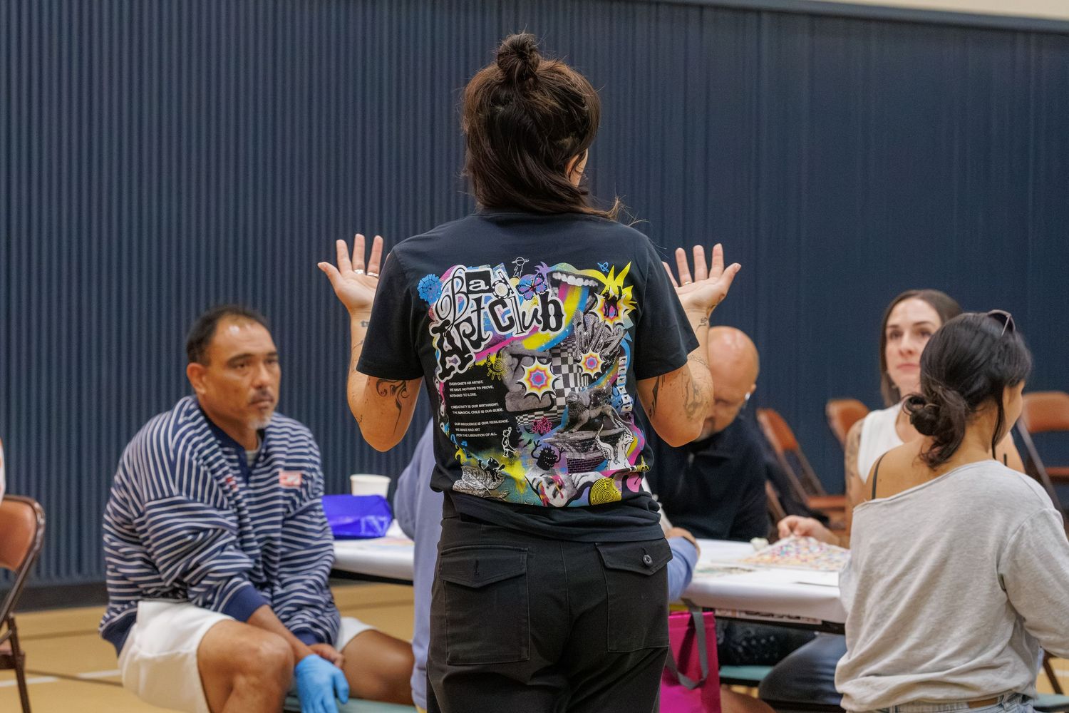 Person in graphic black t-shirt gestures with raised hands to seated group during community health event indoors.