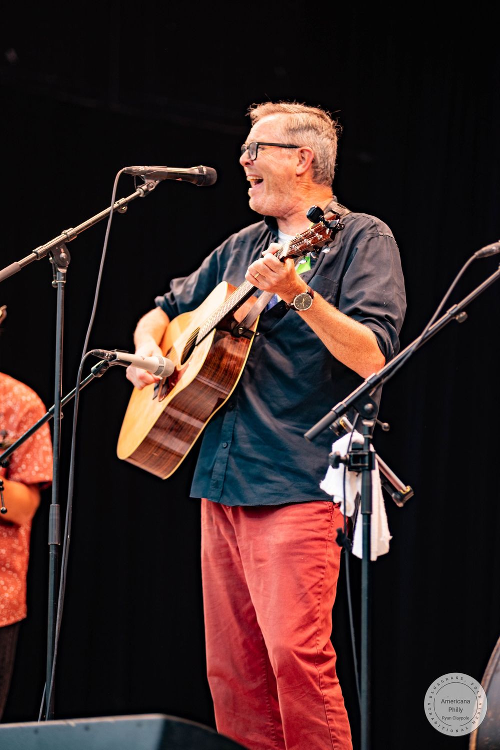Smiling man playing acoustic guitar on stage, wearing red pants and blue shirt with glasses at outdoor music festival.