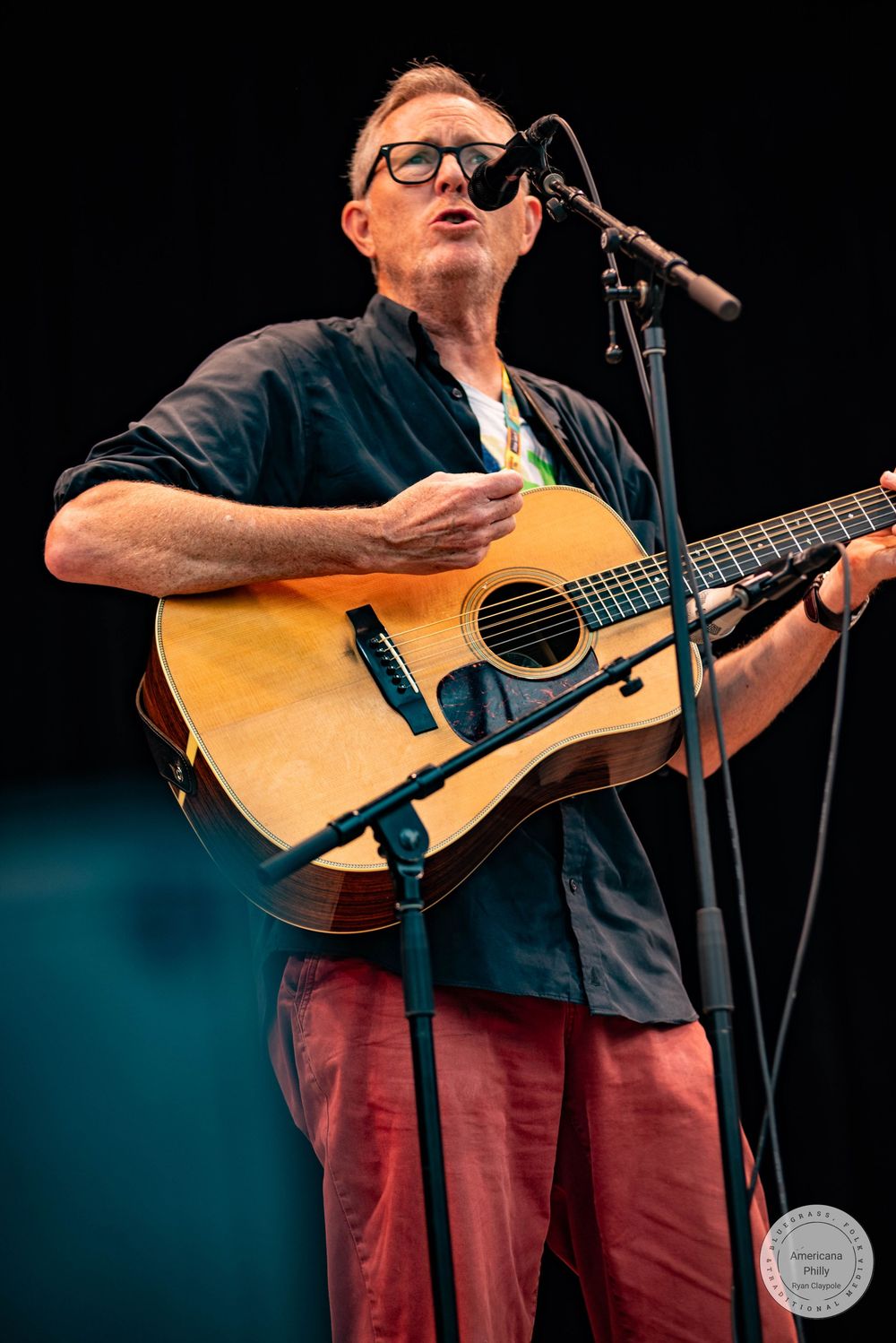 Man playing acoustic guitar and singing into microphone on stage, wearing glasses, dark shirt, and red pants.