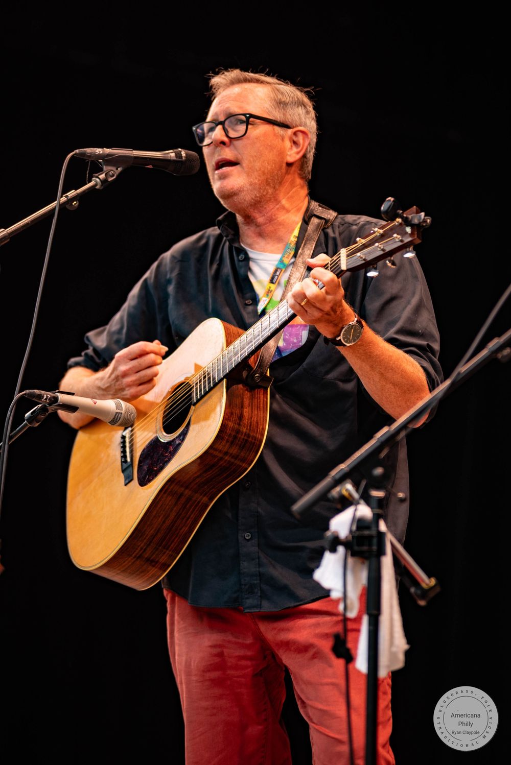 Man in red pants plays acoustic guitar and sings into microphone on stage.