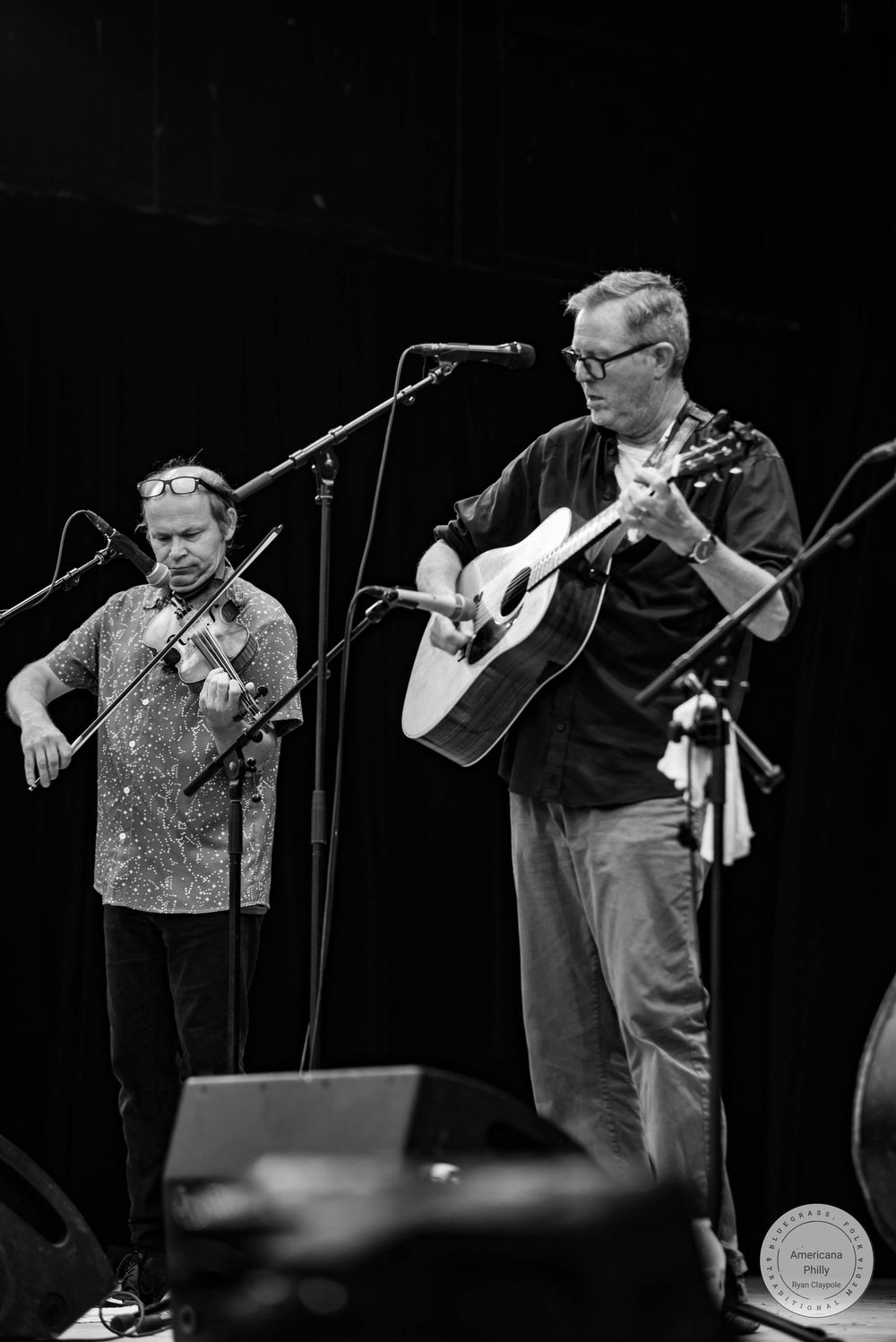 Two musicians perform on stage, one playing violin and one playing acoustic guitar, in a black and white concert photo.