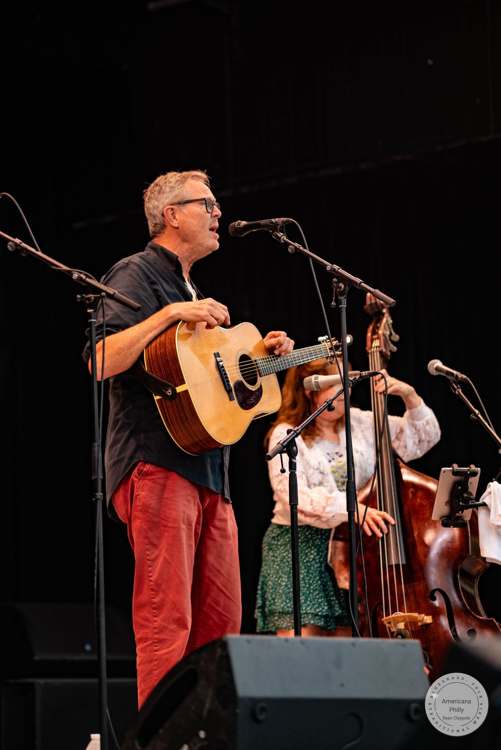 Male guitarist in red pants performs on stage with female bassist behind him.