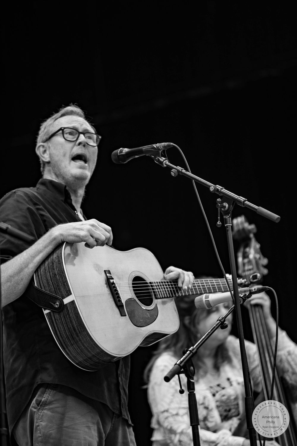 Black and white photo of a man playing acoustic guitar and singing into a microphone on stage.