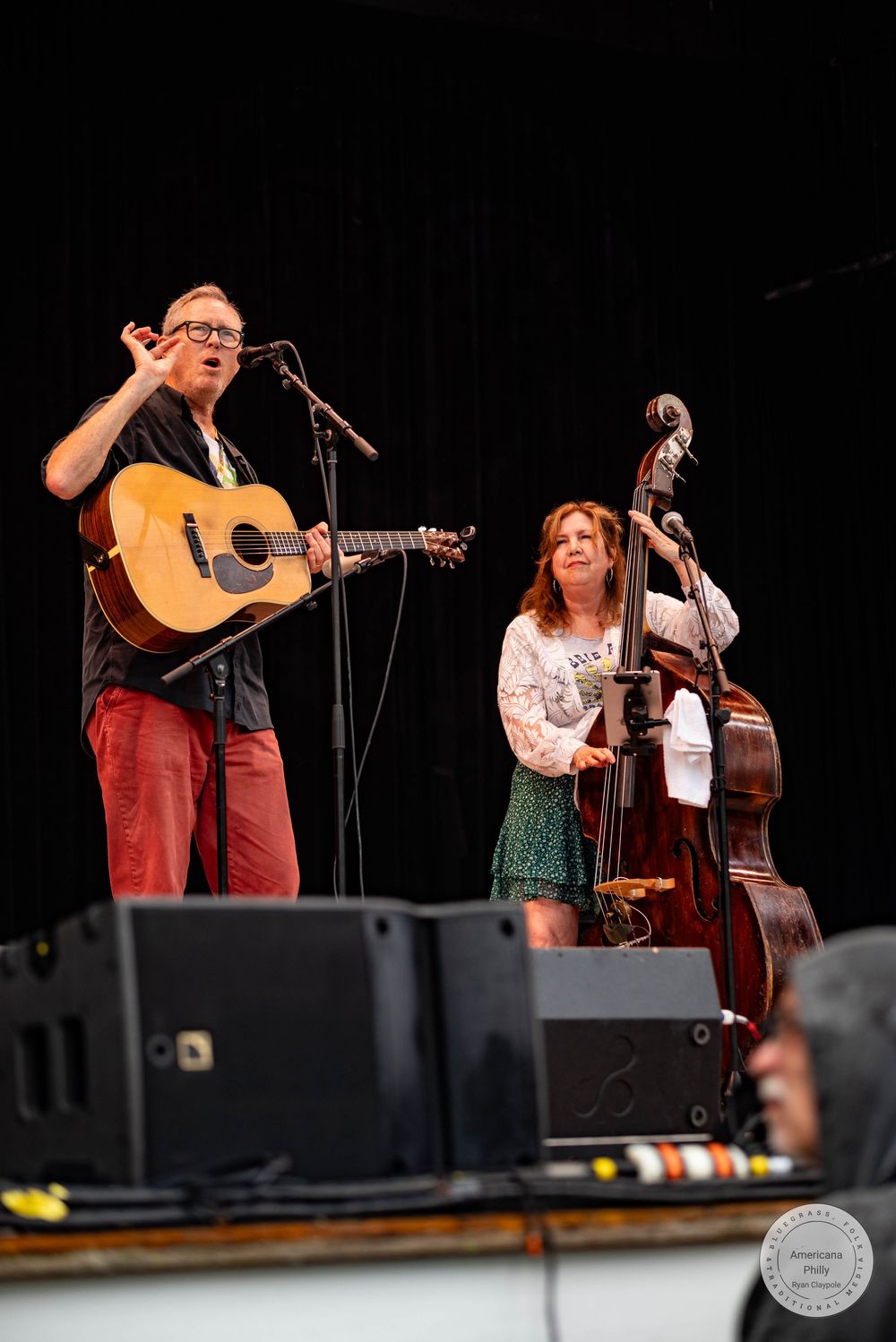 Two musicians perform on stage, one playing acoustic guitar and singing, the other standing beside a upright bass.