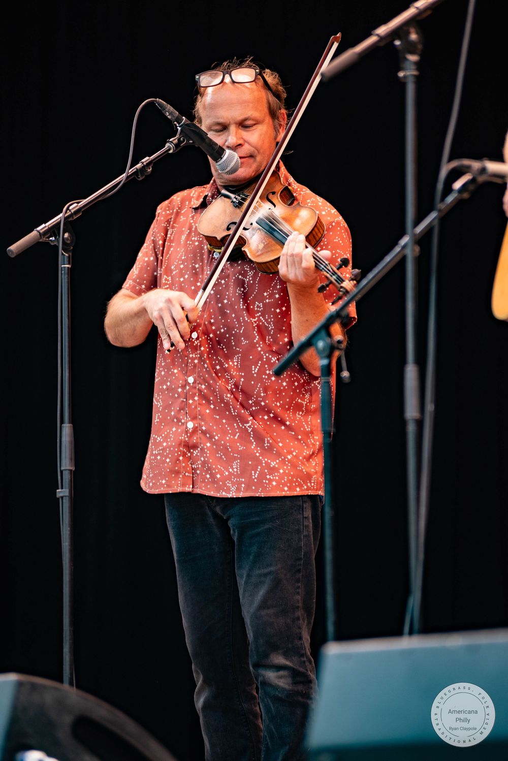 A musician in a red patterned shirt plays violin on stage near a microphone at an outdoor performance.