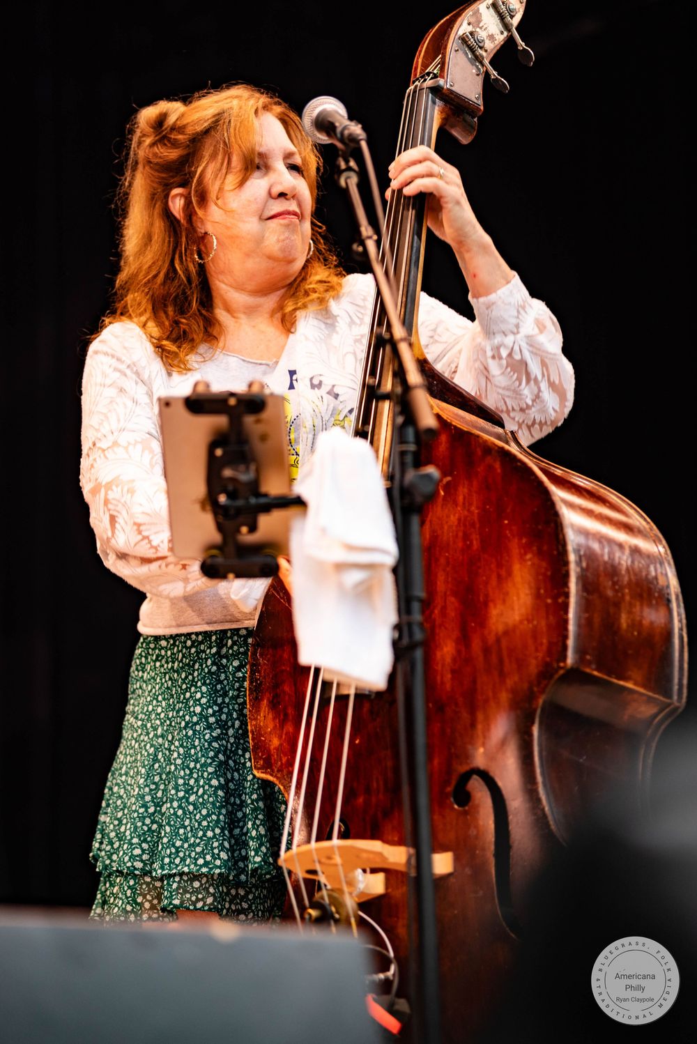 A woman with red hair plays a large upright bass on stage, wearing a white lace top and green floral skirt.