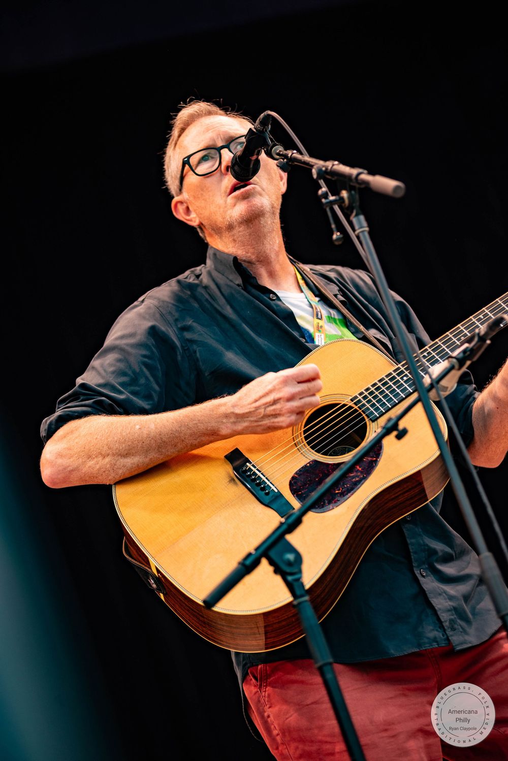A musician with glasses plays acoustic guitar and sings into a microphone on stage wearing a dark shirt and red pants.