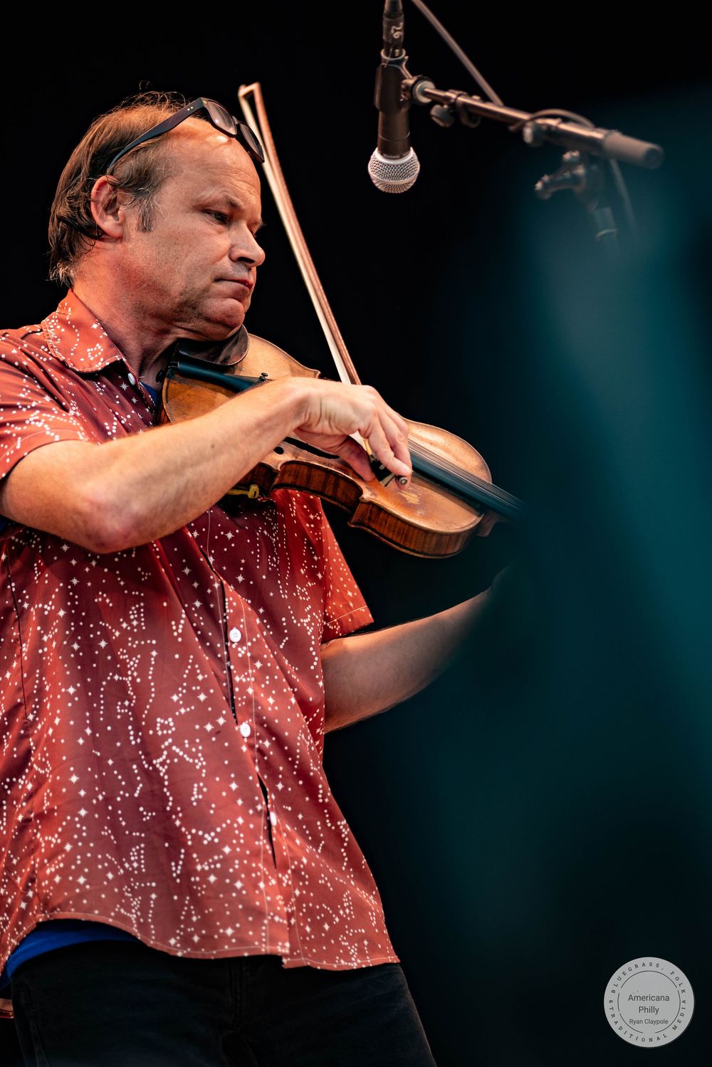 Musician playing violin on stage wearing a red patterned shirt with a bow tucked behind his ear.