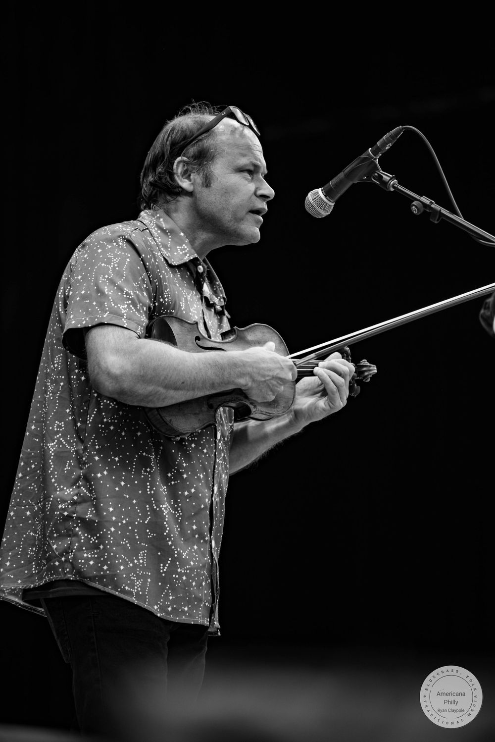 Black and white photo of a man playing violin at a microphone stand during a live performance.