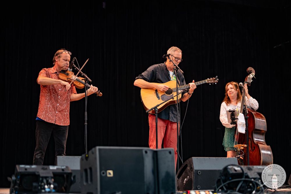 Three musicians perform on stage, playing violin, acoustic guitar, and upright bass at an outdoor folk music festival.