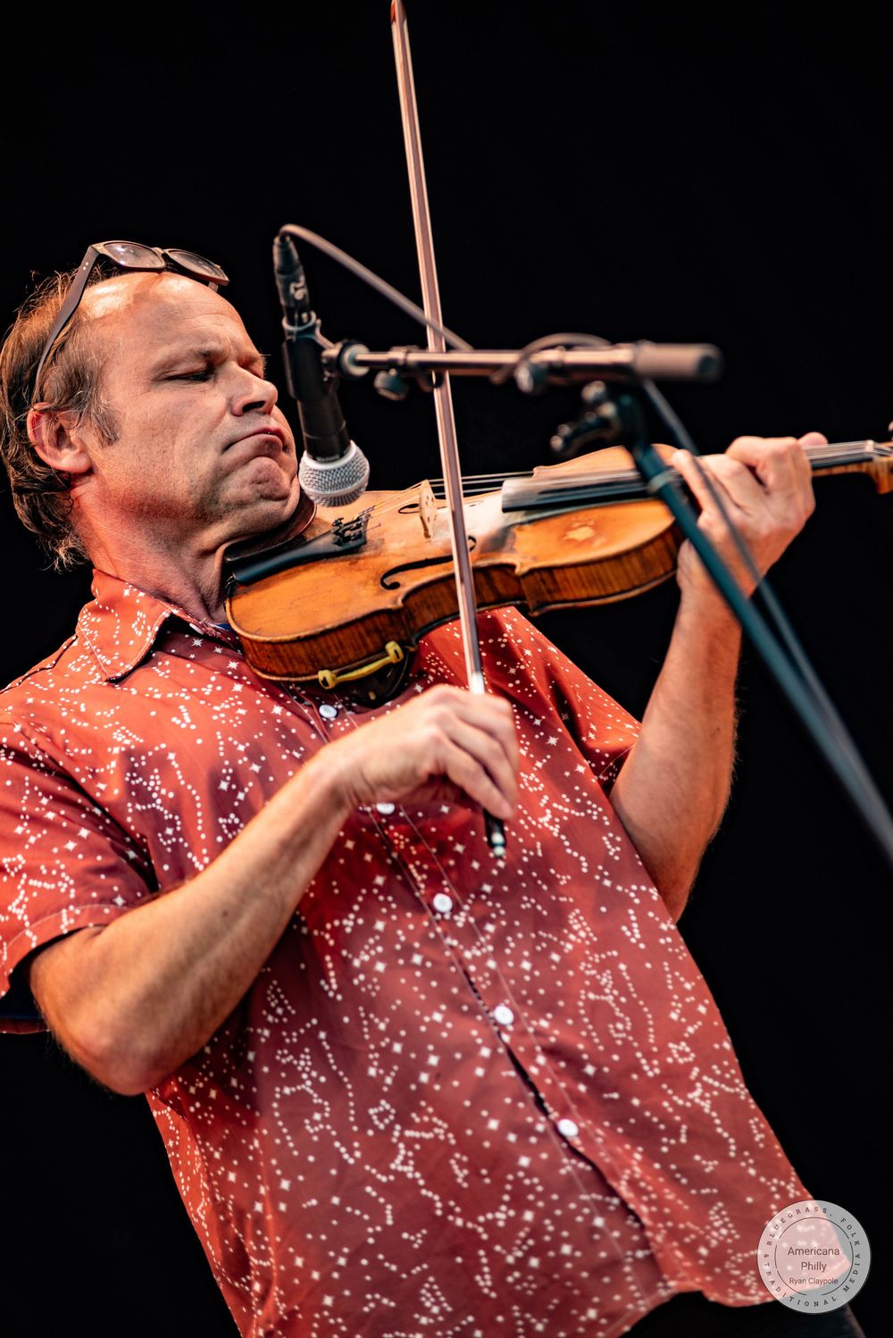 Man in red patterned shirt playing violin on stage at outdoor music festival.