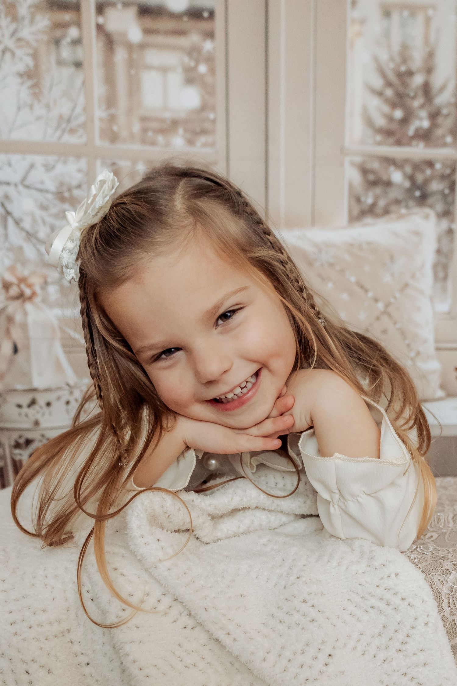 Smiling young girl with braids and white bow rests chin on hands, wrapped in cozy knit blanket near snowy window.