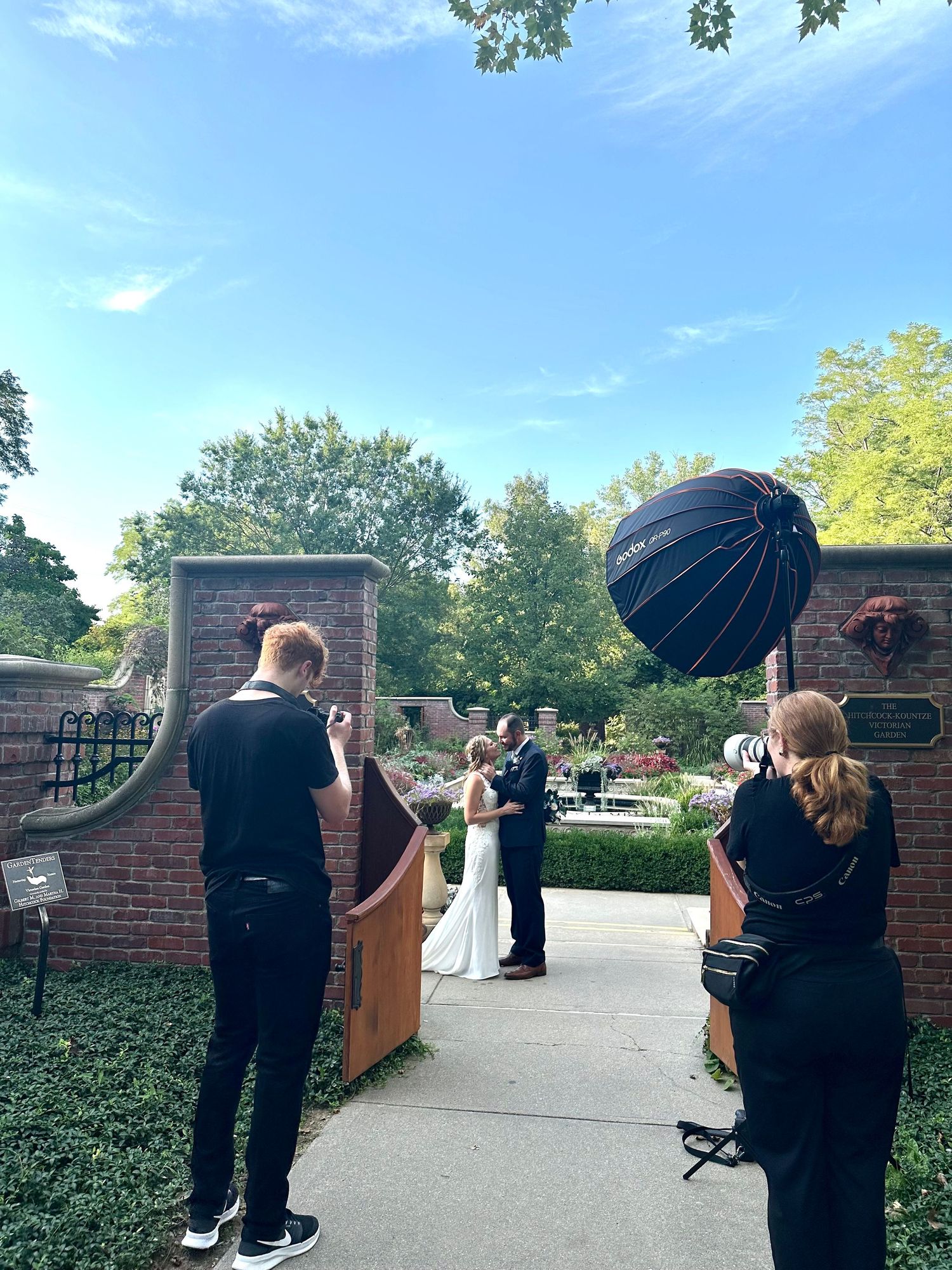 Wedding photographers capturing bride and groom portraits at a brick garden gate entrance on a sunny day.