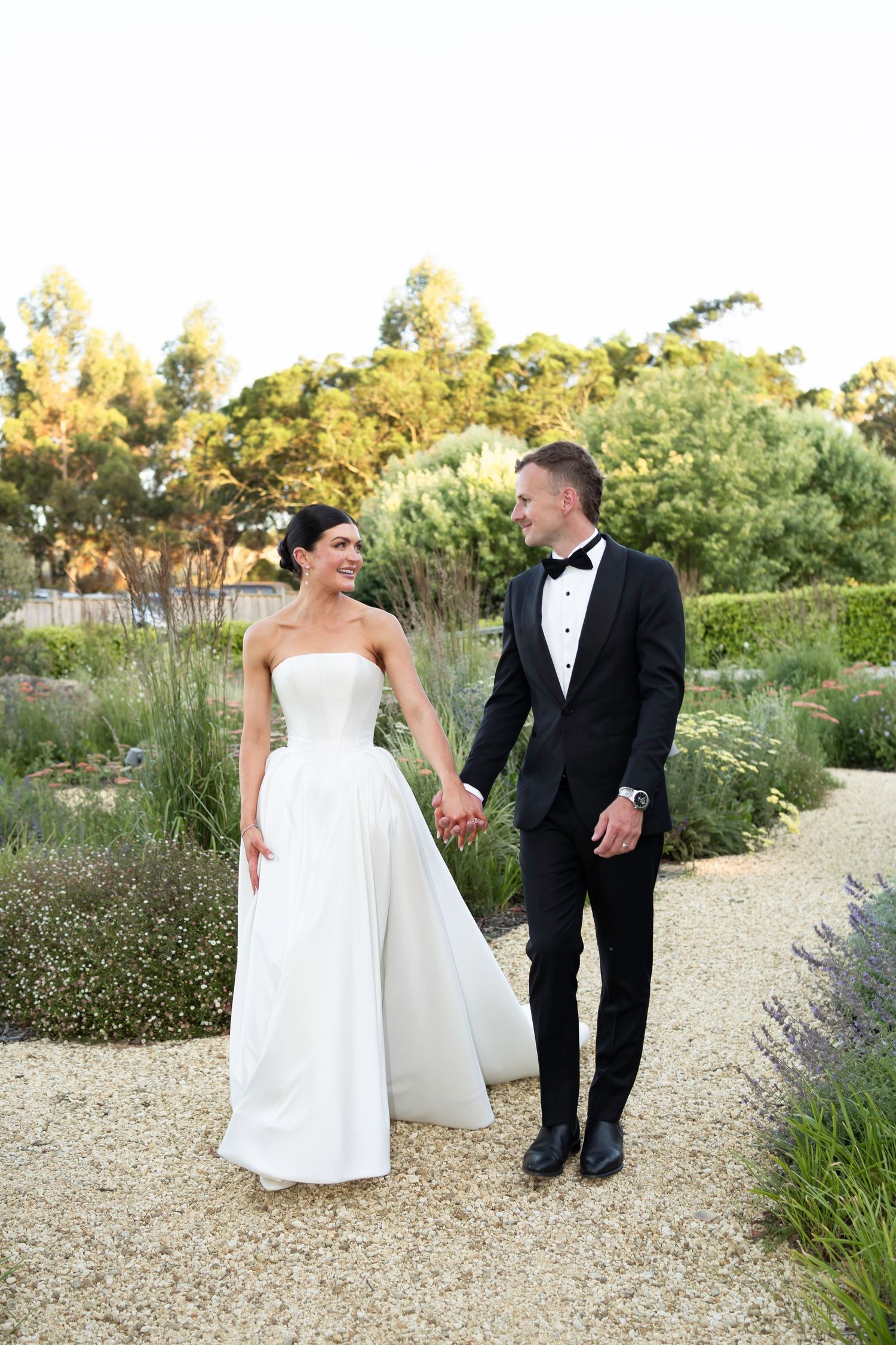A couple in wedding attire walks together on a gravel path surrounded by blooming lavender and garden foliage.