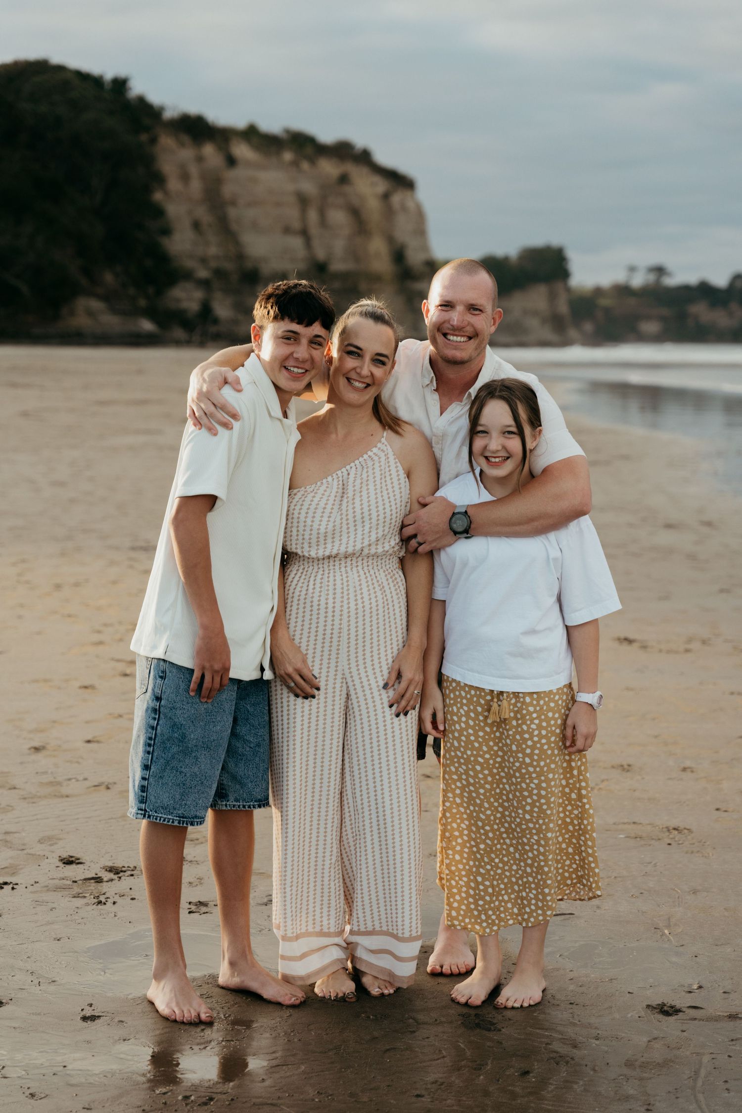 Family of four smiling together on a sandy beach at sunset, with rocky cliffs in the background.