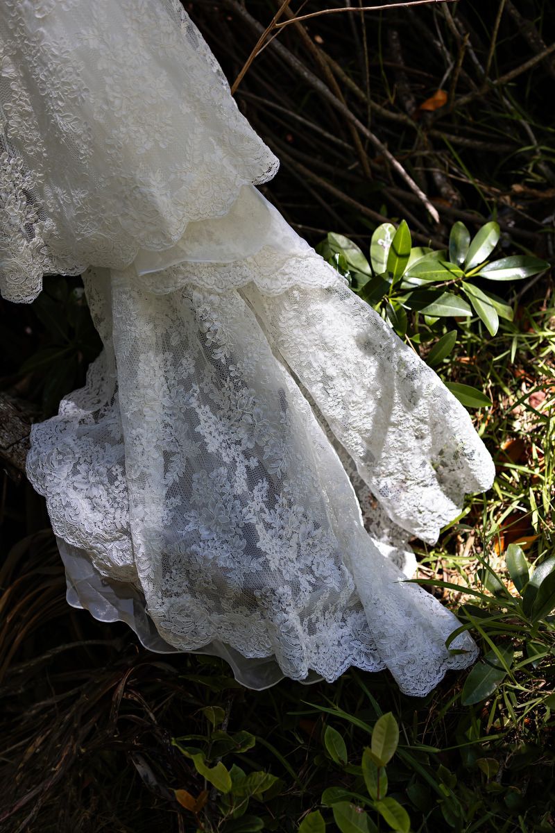 White lace wedding dress train draped over rocks surrounded by green foliage in natural outdoor setting.