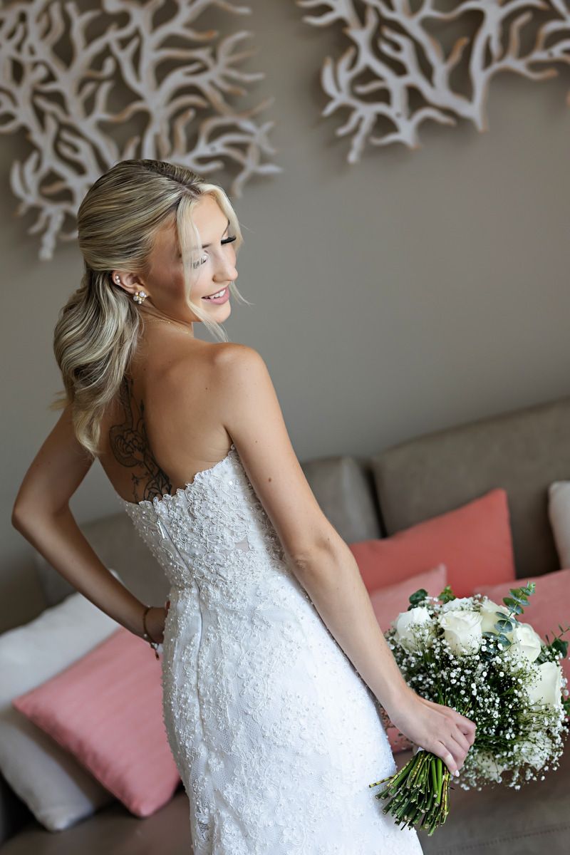 Bride in strapless lace wedding gown holds white bouquet, smiling over shoulder with elegant half-up hairstyle.