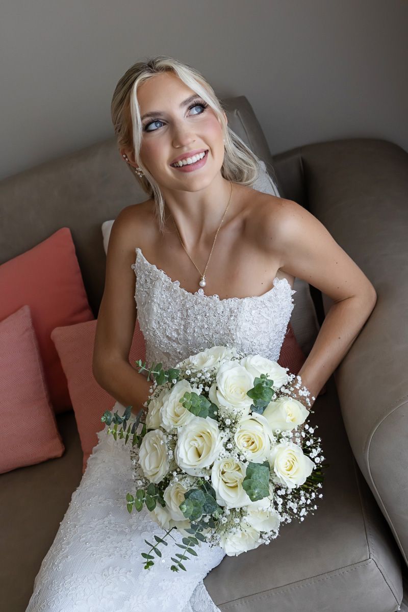 Smiling blonde bride in white lace gown holding white rose bouquet with greenery, seated on beige sofa.