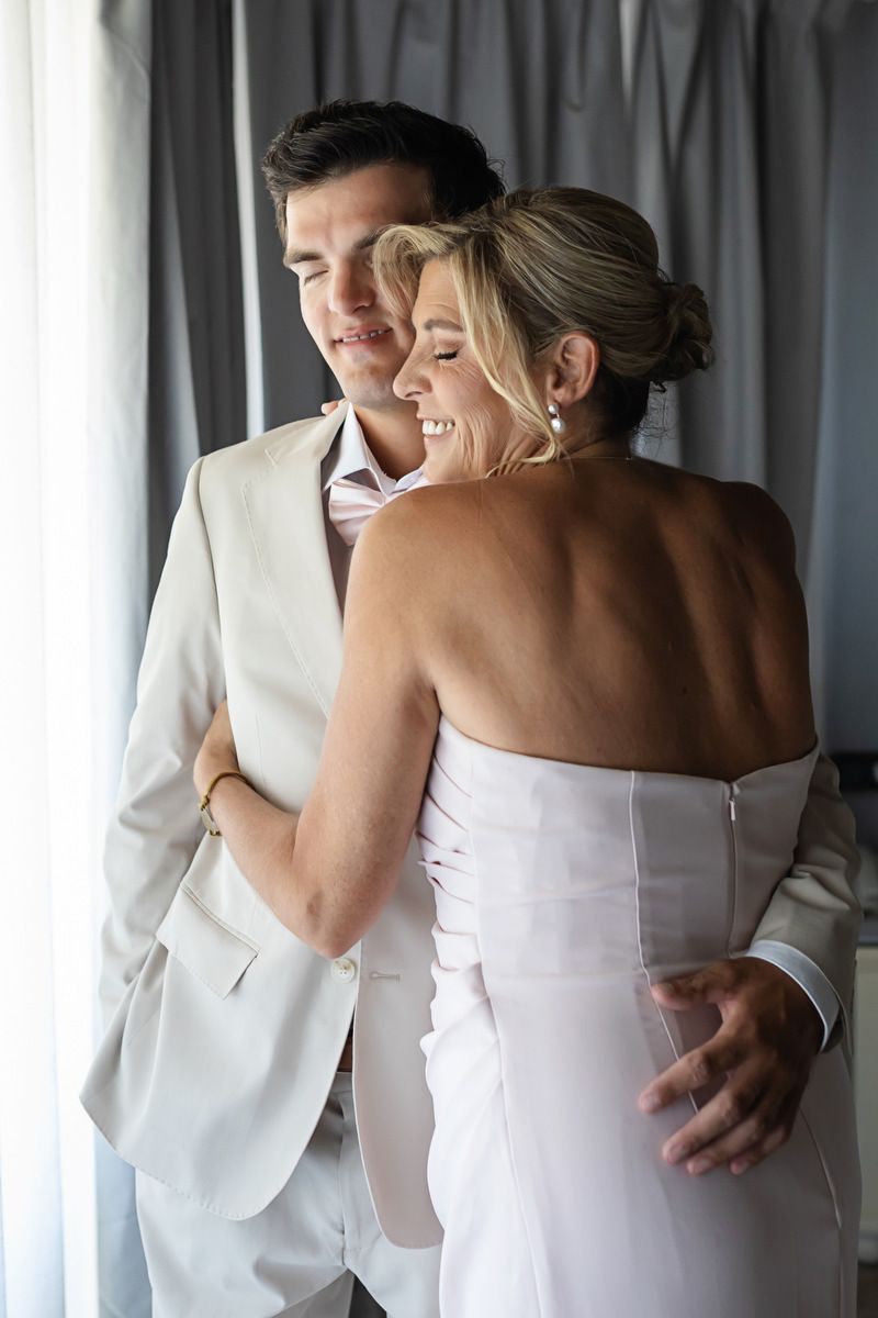 A smiling bride in a white strapless gown embraces a groom in a light suit, sharing a romantic moment indoors.