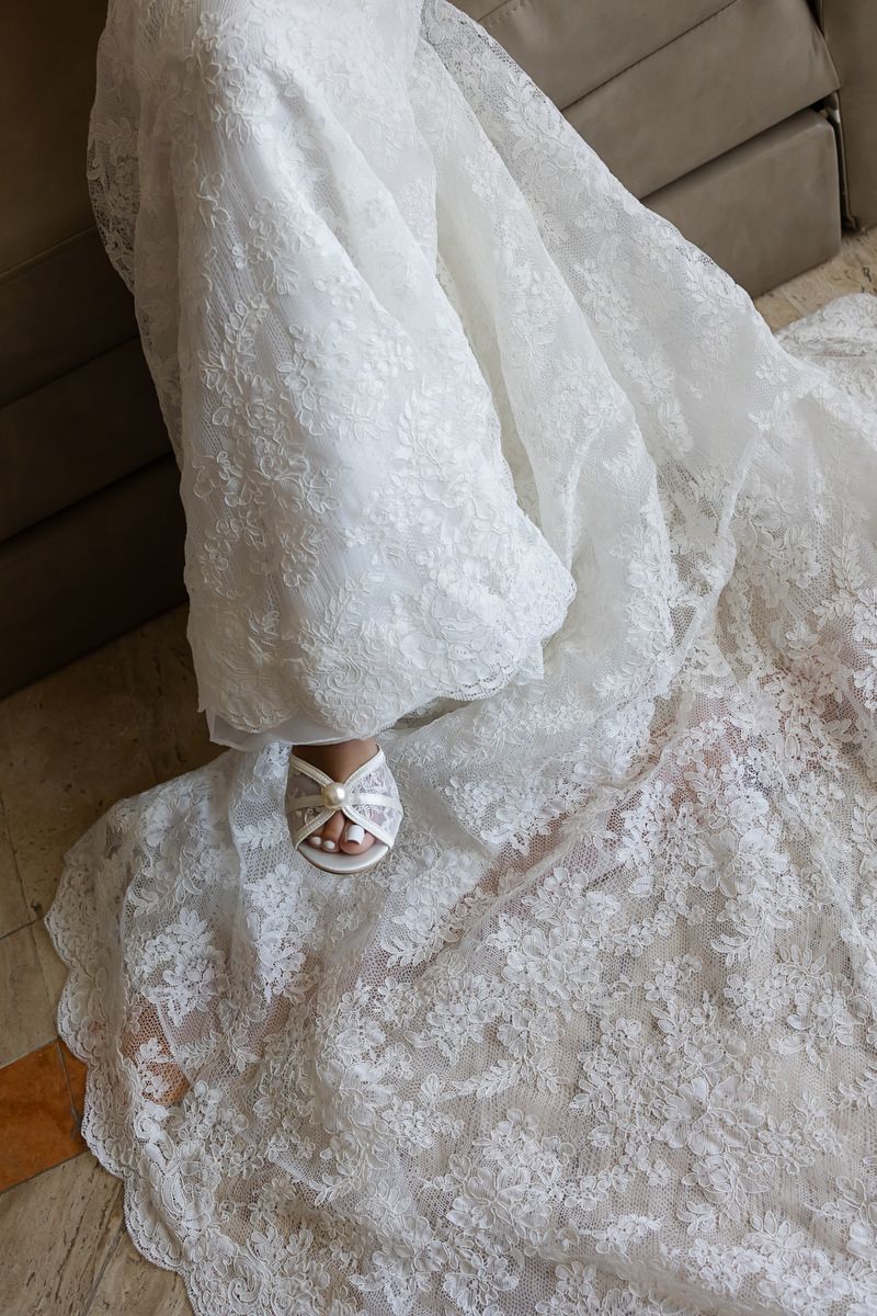 Elegant white lace wedding dress with intricate floral patterns, flowing train, and bridal heels peeking beneath the gown.